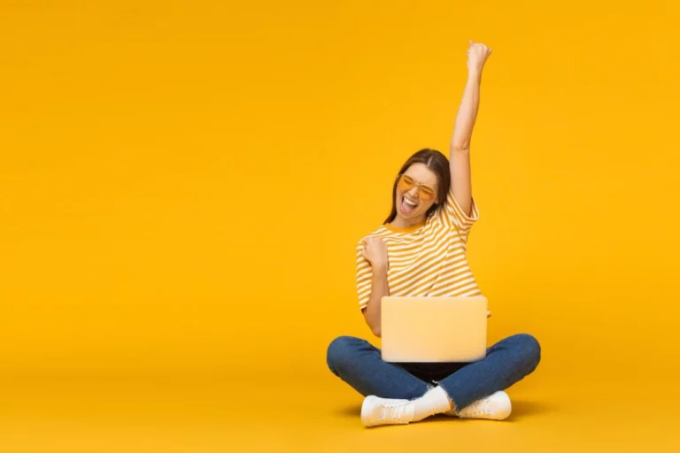 Eine Frau auf orangenem Hintergrund freut sich im Schneidersitz mit einem Laptop auf dem Schoß